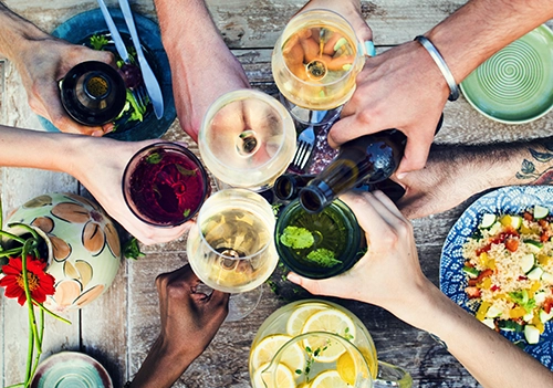 Eine freudige Gruppe junger Menschen stößt mit Wein an, alle sitzen um einen Tisch im Garten bei gemütlichem Sonnenschein.